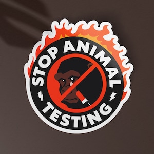 Op de afbeelding: Een rode en witte sticker met een zwarte cirkel in het midden. De cirkel heeft een rode streep erdoorheen en een zwart en bruin apengezicht erin. De tekst "STOP ANIMAL TESTING" staat in witte letters rond de cirkel.
