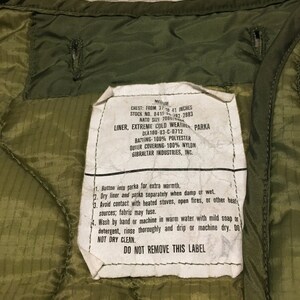 K&ouml;nnte beinhalten: Ein gr&uuml;nes Etikett f&uuml;r eine Milit&auml;rparka-Innenfutter mit Pflege- und Gebrauchsanweisungen. Das Etikett lautet "Chest: From 37 to 41 inches", "Stock No. 8415-082-2883", "NATO Size 7080101", "Liner, Extreme Cold Weather Parka", "DIA 100-83-C-0712", "Batting-100% Polyester", "Outer Covering-100% Nylon", "Gibraltar Industries, Inc.", und "Do Not Remove This Label".