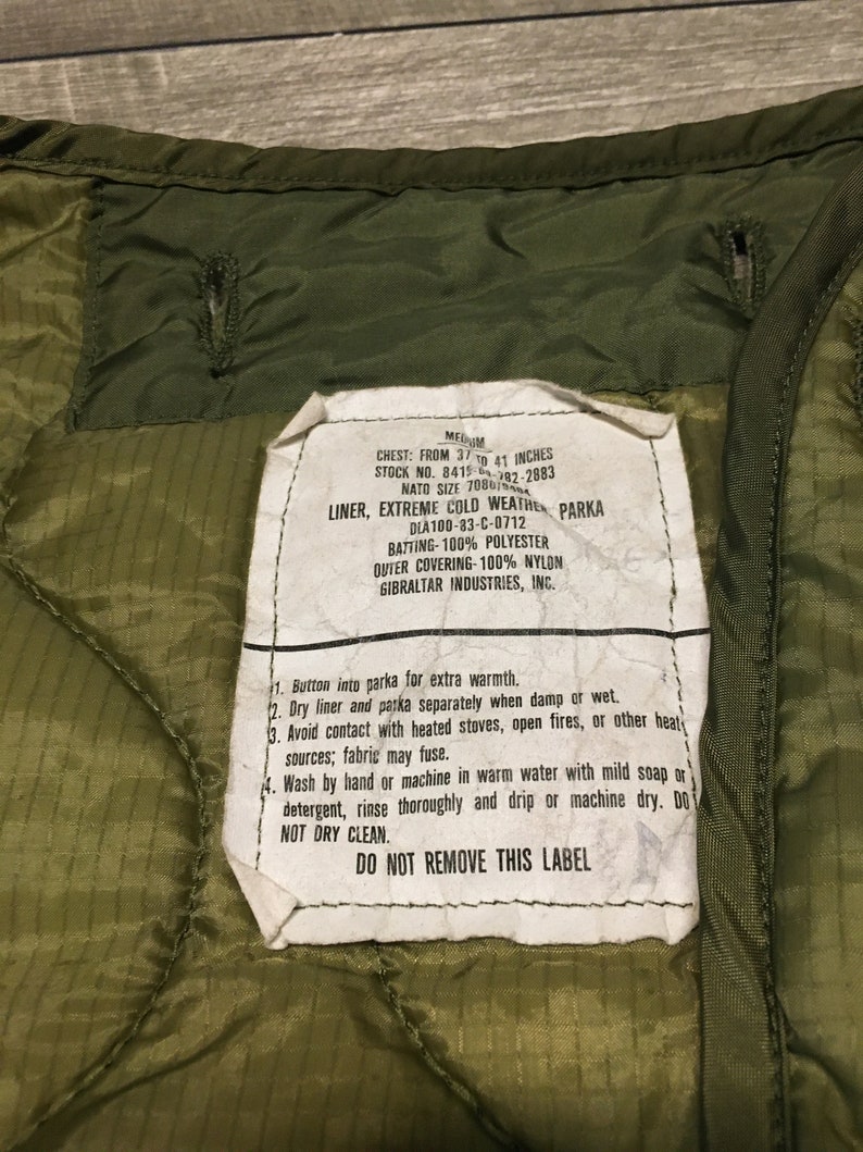 K&ouml;nnte beinhalten: Ein gr&uuml;nes Etikett f&uuml;r eine Milit&auml;rparka-Innenfutter mit Pflege- und Gebrauchsanweisungen. Das Etikett lautet "Chest: From 37 to 41 inches", "Stock No. 8415-082-2883", "NATO Size 7080101", "Liner, Extreme Cold Weather Parka", "DIA 100-83-C-0712", "Batting-100% Polyester", "Outer Covering-100% Nylon", "Gibraltar Industries, Inc.", und "Do Not Remove This Label".