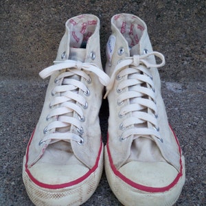 Pode incluir: Um par de t&ecirc;nis branco de lona de cano alto com acabamento vermelho ao redor da sola. Os cadar&ccedil;os s&atilde;o brancos e os t&ecirc;nis t&ecirc;m um visual vintage.