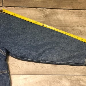 May include: A blue denim jacket sleeve with a visible yellow measuring tape placed along the top, showing a length of approximately 27 inches from the shoulder seam to the cuff. The jacket features a chest pocket and visible stitching details.