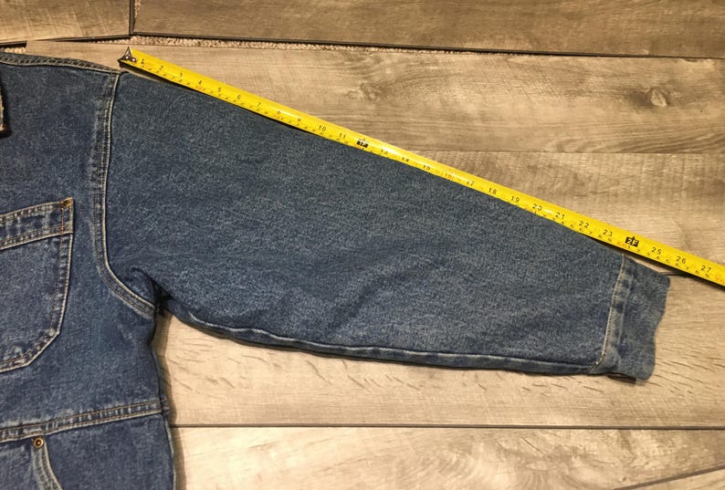 May include: A blue denim jacket sleeve with a visible yellow measuring tape placed along the top, showing a length of approximately 27 inches from the shoulder seam to the cuff. The jacket features a chest pocket and visible stitching details.