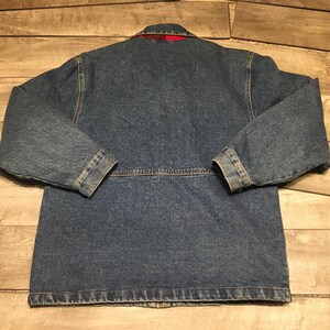 May include: A blue denim jacket featuring long sleeves, a collar, and a horizontal seam across the back. The interior lining is visible at the collar and displays a red and black plaid pattern. The jacket is constructed with visible stitching.
