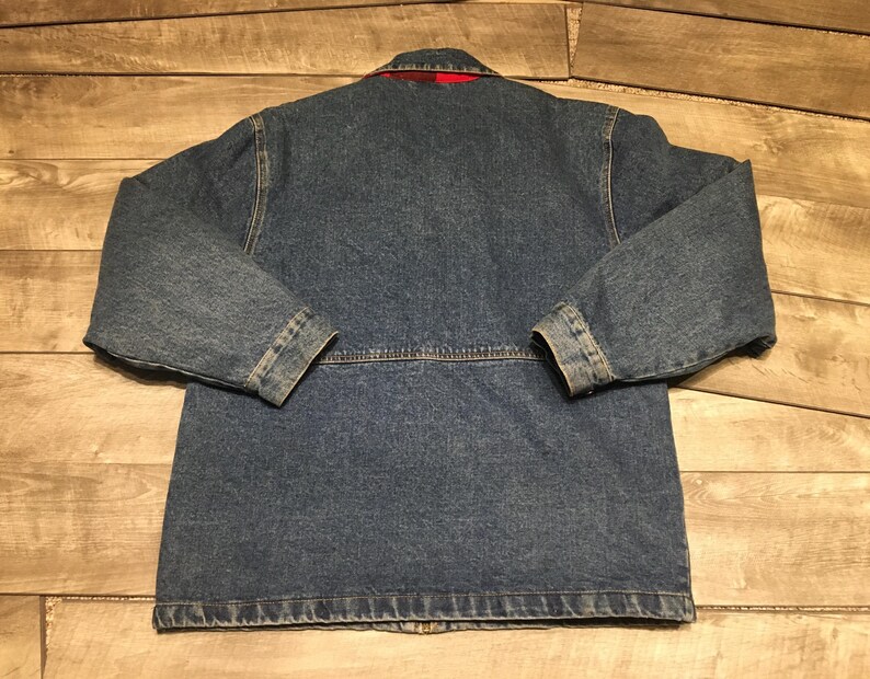 May include: A blue denim jacket featuring long sleeves, a collar, and a horizontal seam across the back. The interior lining is visible at the collar and displays a red and black plaid pattern. The jacket is constructed with visible stitching.