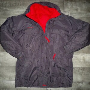 Puede incluir: Una chaqueta gris oscuro con forro de polar rojo. La chaqueta tiene cierre de cremallera y botones, dos bolsillos en la parte delantera y un cordón en la cintura.