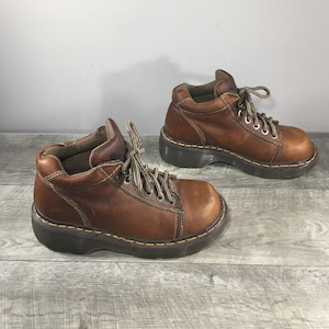 Può includere: Un paio di stivaletti in pelle marrone con lacci. Gli stivaletti hanno la punta arrotondata, una suola spessa e cuciture a vista. I lacci sono marroni e beige. Gli stivaletti misurano circa 15 cm di lunghezza.