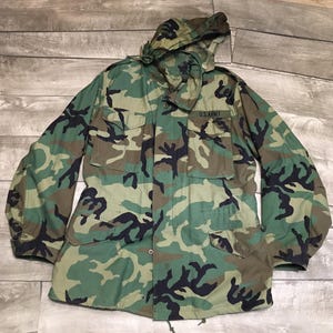 May include: A camouflage pattern jacket with a hood. The jacket features a green, brown, and black woodland camouflage design. The text "U.S. ARMY" is visible on the upper right chest. The jacket has multiple pockets and a button-up front.