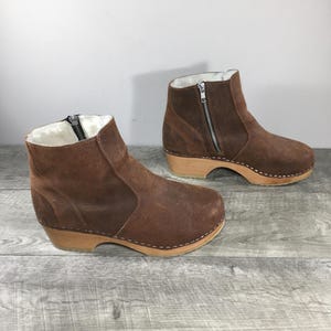 Pode incluir: Duas botas de tornozelo de camurça castanha com solas de madeira. As botas têm fecho de correr lateral e forro de pele sintética. As solas são de madeira clara e são presas com pequenas costuras brancas.