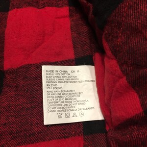 May include: A red and black buffalo plaid quilted fabric with a white care label attached. The label text reads: MADE IN CHINA CM 11, SHELL: 100% COTTON, BODY LINING: 100% COTTON, SLEEVE LINING: 100% NYLON, PADDING: 100% POLYESTER RESIN TREATED, RN 23923, P.O. #50835.