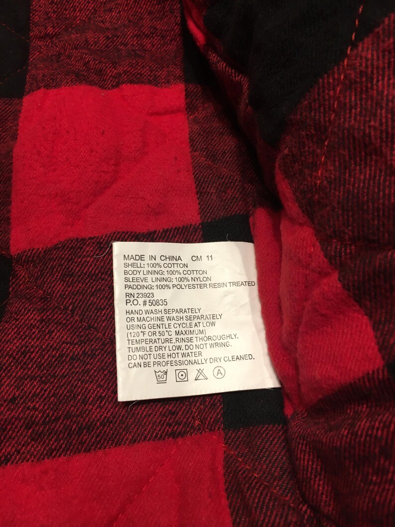 May include: A red and black buffalo plaid quilted fabric with a white care label attached. The label text reads: MADE IN CHINA CM 11, SHELL: 100% COTTON, BODY LINING: 100% COTTON, SLEEVE LINING: 100% NYLON, PADDING: 100% POLYESTER RESIN TREATED, RN 23923, P.O. #50835.