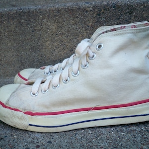 Pode incluir: T&ecirc;nis branco de lona de cano alto com acabamento vermelho e azul ao redor da sola. Os cadar&ccedil;os s&atilde;o brancos e os t&ecirc;nis est&atilde;o desgastados.