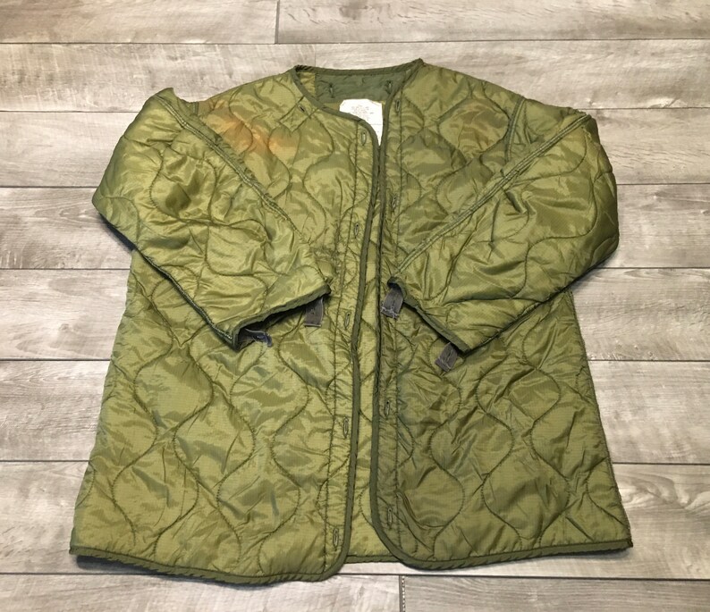 K&ouml;nnte beinhalten: Eine gr&uuml;ne gesteppte Jacke mit Druckknopfverschluss. Die Jacke hat ein Rautenmuster und ist aus leichtem Material gefertigt.