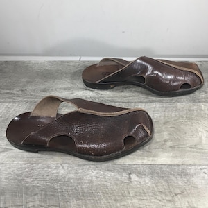 May include: Two brown leather sandals with open toes and a strap across the top. The sandals have a textured appearance and are set against a light gray wooden surface.