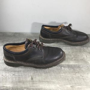 Può includere: Un paio di scarpe Oxford in pelle marrone con lacci marrone scuro. Le scarpe hanno una punta arrotondata e una suola spessa e strutturata. L'interno è beige con un bordo blu attorno all'apertura. Le scarpe sono appoggiate su una superficie di legno grigio.