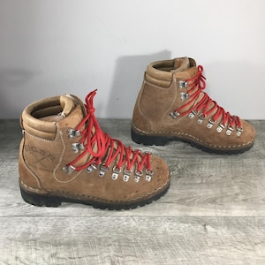 Puede incluir: Un par de botas de senderismo de ante marrón con cordones rojos. Las botas tienen ojales metálicos y una suela negra gruesa. Las botas están etiquetadas "Eiger" en el lateral. Probablemente son vintage.