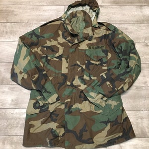 K&ouml;nnte beinhalten: Eine gr&uuml;ne, braune und schwarze Tarnjacke mit Kapuze und Knopfleiste. Die Jacke hat einen Aufn&auml;her auf der linken Brust mit der Aufschrift "U.S. ARMY".