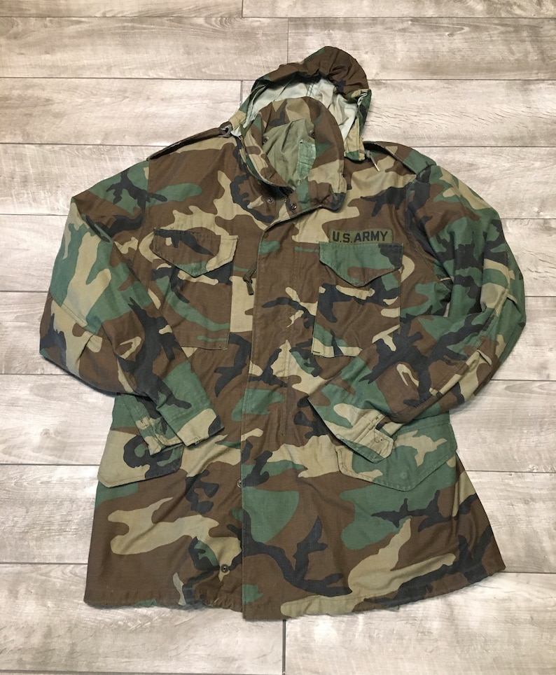 K&ouml;nnte beinhalten: Eine gr&uuml;ne, braune und schwarze Tarnjacke mit Kapuze und Knopfleiste. Die Jacke hat einen Aufn&auml;her auf der linken Brust mit der Aufschrift "U.S. ARMY".