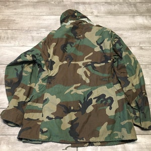 K&ouml;nnte beinhalten: Eine gr&uuml;ne, braune und schwarze Tarnjacke mit Kapuze. Die Jacke hat einen Rei&szlig;verschluss und mehrere Taschen.