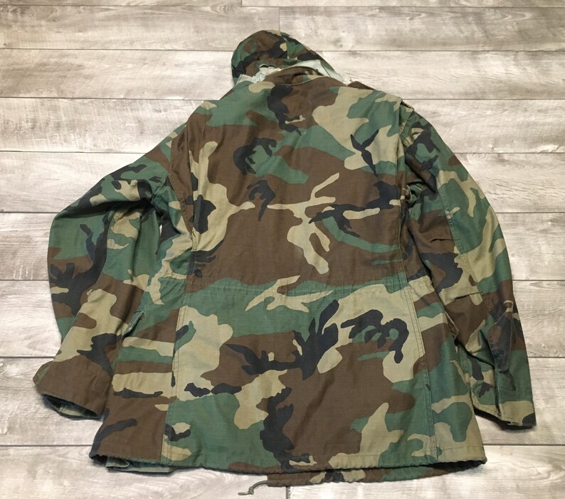 K&ouml;nnte beinhalten: Eine gr&uuml;ne, braune und schwarze Tarnjacke mit Kapuze. Die Jacke hat einen Rei&szlig;verschluss und mehrere Taschen.