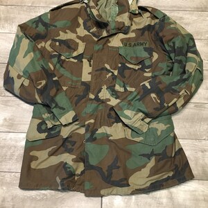 K&ouml;nnte beinhalten: Eine gr&uuml;ne, braune und schwarze Tarnjacke mit einem U.S. Army-Aufn&auml;her auf der linken Brust.