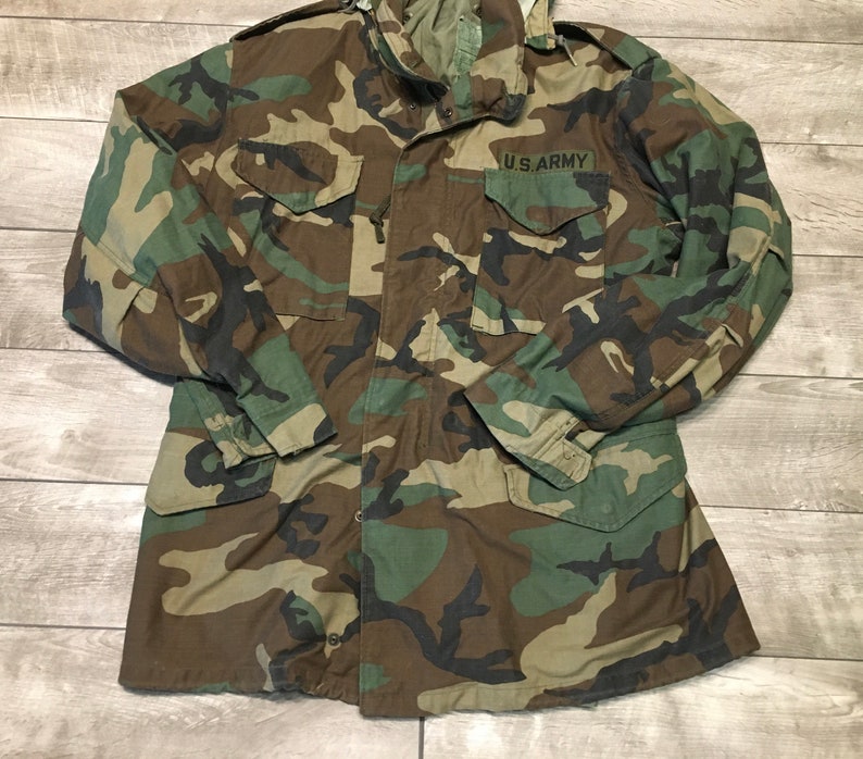 K&ouml;nnte beinhalten: Eine gr&uuml;ne, braune und schwarze Tarnjacke mit einem U.S. Army-Aufn&auml;her auf der linken Brust.