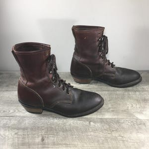 Puede incluir: Un par de botas de cuero marrón oscuro con cordones, altura media, puntera redondeada y tacón de madera apilada. Las botas tienen cordones marrones a juego en ojales metálicos y costuras visibles a lo largo de los paneles laterales y el talón.