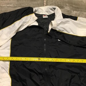 May include: Black, white, and yellow Nike track jacket. The jacket has a full zipper, a white collar, and white sleeves with yellow trim. A yellow measuring tape is laid across the jacket, showing a measurement of approximately 27 inches.