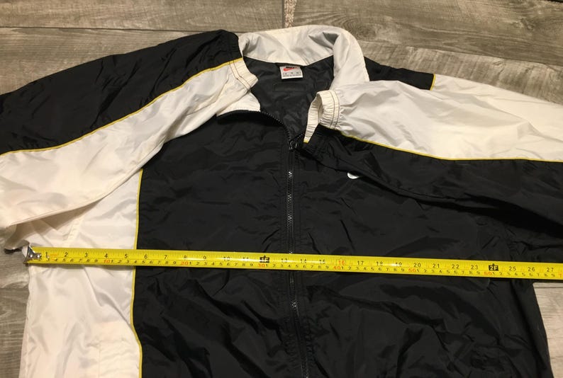 May include: Black, white, and yellow Nike track jacket. The jacket has a full zipper, a white collar, and white sleeves with yellow trim. A yellow measuring tape is laid across the jacket, showing a measurement of approximately 27 inches.