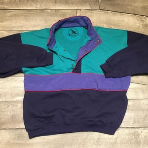Pode incluir: Um moletom de gola alta com blocos de cores em verde-azulado, roxo e azul marinho. O moletom tem gola abotoada e mangas compridas. Uma borda vermelha acentua a seção roxa. A gola é roxa com o interior verde-azulado.