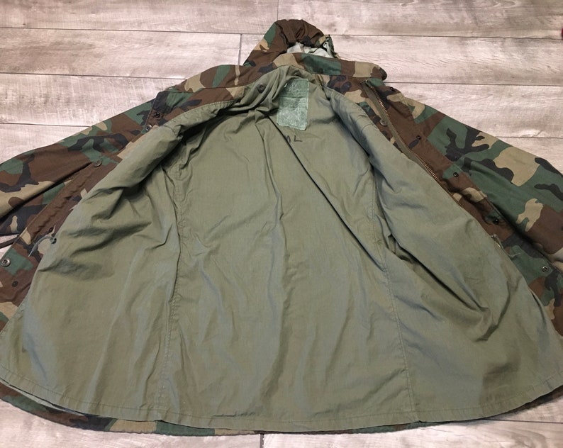 K&ouml;nnte beinhalten: Eine gr&uuml;ne, braune und schwarze Tarnjacke mit Kapuze. Die Jacke ist ge&ouml;ffnet und auf links gedreht, wodurch ein gr&uuml;nes Futter sichtbar wird.