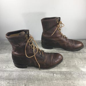 Puede incluir: Un par de botas de cuero marrón oscuro con cordones, diseño de caña alta, punteras redondeadas y ojales metálicos en tono latón. Las botas tienen cordones color canela, un contrafuerte reforzado y suela oscura, con desgaste visible en el cuero.