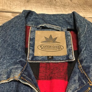 May include: A blue denim jacket with a red and black plaid lining. A tan rectangular patch is stitched to the interior collar area featuring the text Canyon Guide Outfitters with a star icon and the size M printed on a small black tag below.