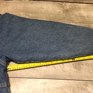 May include: A blue denim jacket sleeve with visible stitching and a cuff, positioned alongside a yellow measuring tape that indicates a length of approximately 20 inches from the shoulder seam to the end of the sleeve cuff.