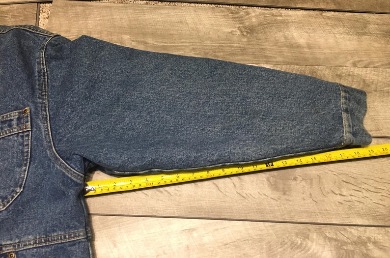 May include: A blue denim jacket sleeve with visible stitching and a cuff, positioned alongside a yellow measuring tape that indicates a length of approximately 20 inches from the shoulder seam to the end of the sleeve cuff.