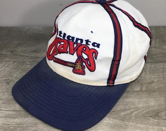 Könnte beinhalten: Weiße Baseballkappe mit einem rot-blauen, gestreiften Band. Die Vorderseite der Kappe zeigt ein rotes und weißes gesticktes Logo mit der Aufschrift "Atlanta Braves" und einer Tomahawk.