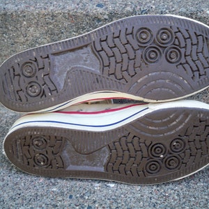 Pode incluir: Close-up da parte inferior de um par de t&ecirc;nis de lona branca com solas de borracha marrom. As solas t&ecirc;m um padr&atilde;o texturizado com reentr&acirc;ncias circulares.