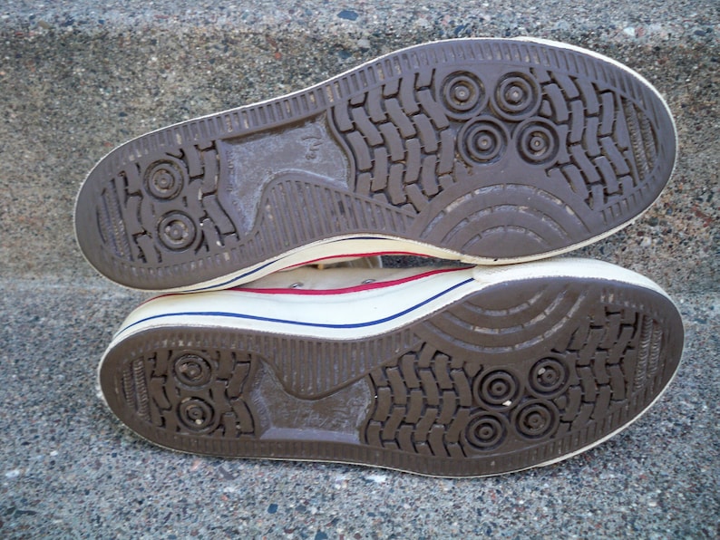 Pode incluir: Close-up da parte inferior de um par de t&ecirc;nis de lona branca com solas de borracha marrom. As solas t&ecirc;m um padr&atilde;o texturizado com reentr&acirc;ncias circulares.