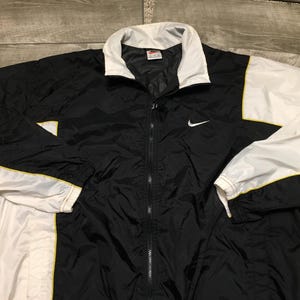 May include: Black and white Nike windbreaker jacket with a full zipper closure. The jacket features a white collar, white sleeves, and yellow trim. A white Nike swoosh logo is on the left chest.