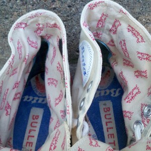 Pode incluir: Um par de t&ecirc;nis branco de lona de cano alto com texto vermelho que diz "Protected" e "Pacifata" no interior do sapato. A palmilha &eacute; azul e diz "Bullet" em letras brancas.