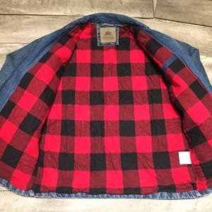 May include: A blue denim jacket featuring a quilted interior lining with a red and black buffalo plaid pattern. The jacket includes a collar, long sleeves, and a rectangular brand label stitched at the center of the inner neck area.