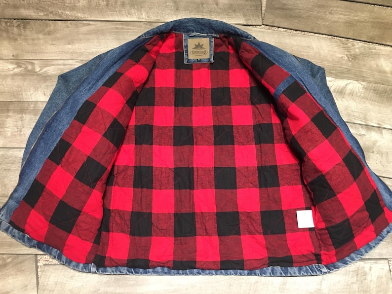 May include: A blue denim jacket featuring a quilted interior lining with a red and black buffalo plaid pattern. The jacket includes a collar, long sleeves, and a rectangular brand label stitched at the center of the inner neck area.