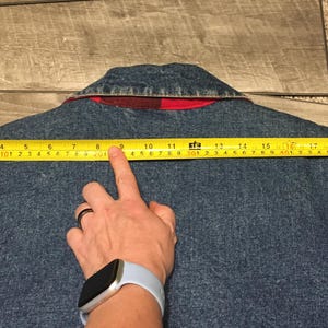 May include: A blue denim jacket with a red plaid inner collar lining. A yellow measuring tape is positioned across the back shoulders, showing a width of approximately 22 inches from the left seam to the right seam of the garment.