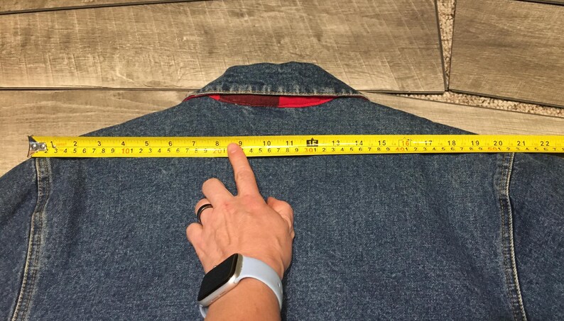 May include: A blue denim jacket with a red plaid inner collar lining. A yellow measuring tape is positioned across the back shoulders, showing a width of approximately 22 inches from the left seam to the right seam of the garment.