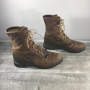 Puede incluir: Un par de botas de cuero marrón con cordones, altura media, puntera redonda y un detalle de flecos decorativos sobre la lengüeta. Las botas tienen ojales de color latón, cordones color canela y una suela de goma marrón oscuro.