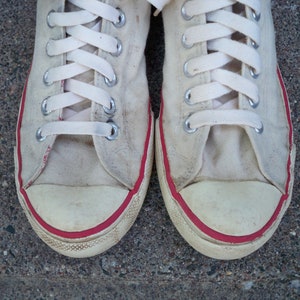 Pode incluir: Um par de t&ecirc;nis branco de lona com acabamento vermelho ao redor das bordas. Os cadar&ccedil;os s&atilde;o brancos e os t&ecirc;nis est&atilde;o desgastados.