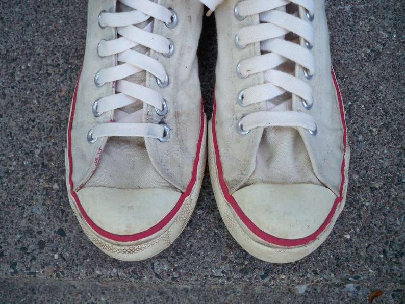 Pode incluir: Um par de t&ecirc;nis branco de lona com acabamento vermelho ao redor das bordas. Os cadar&ccedil;os s&atilde;o brancos e os t&ecirc;nis est&atilde;o desgastados.