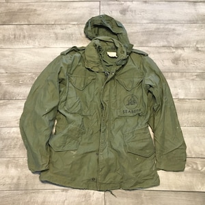 May include: A green military jacket with a hood. The jacket has a patch on the left chest that says "Seabees".