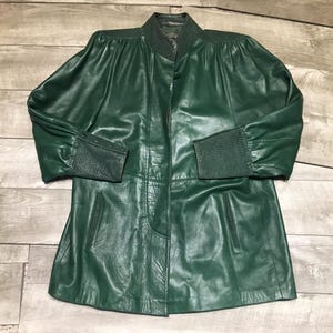 May include: A dark green leather jacket with a button closure and a collared neckline. The jacket has long sleeves with textured cuffs and a tailored fit. The jacket is approximately 36 inches long.
