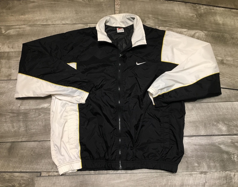 May include: Black and white Nike track jacket with a white collar and sleeves. The jacket features a yellow stripe detail and a small Nike swoosh logo on the chest. The jacket is fully zipped and appears to be made of a lightweight material.
