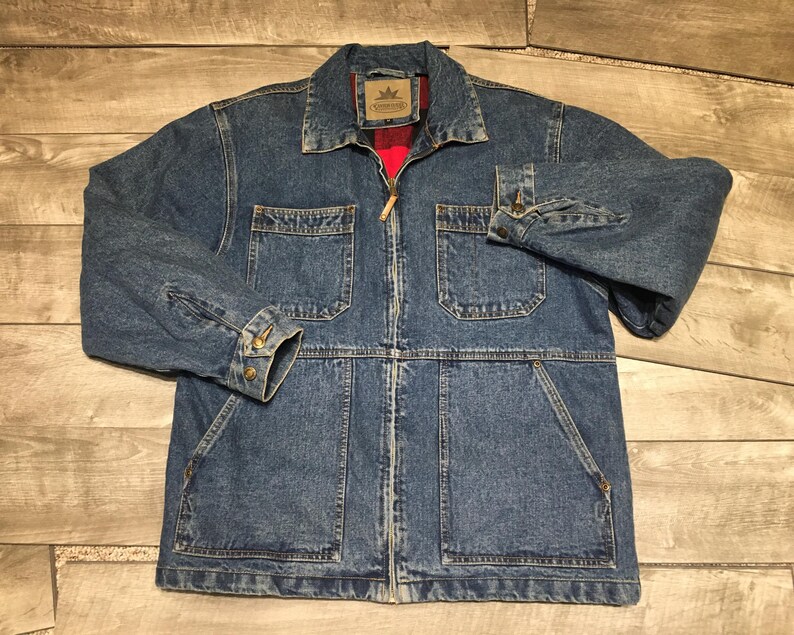 May include: A blue denim jacket featuring a full-length front zipper, a pointed collar, and long sleeves with buttoned cuffs. The jacket includes two chest pockets with flaps and two large lower pockets. A red lining is visible inside the collar.